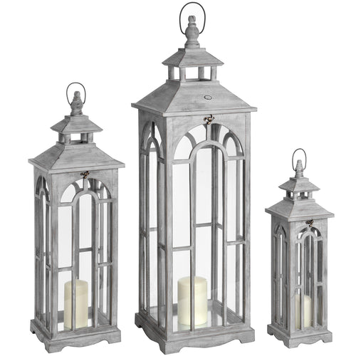 Set of three wooden lanterns with white-wash finish and archway design, each with an aged brass clasp and containing a white candle. They are displayed in a递减的规模.