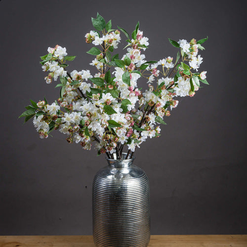 A lifelike faux floral stem with pale pink Prunus blossoms and vibrant green leaves in a silver vase against a dark background.