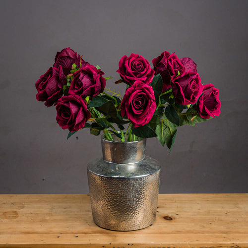 A bouquet of artificial deep red roses arranged in a silver metallic vase, placed on a wooden surface against a gray background.