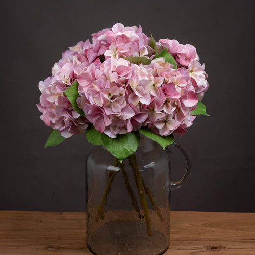 A shabby pink hydrangea artificial flower arrangement placed in a clear glass vase, set against a dark background.