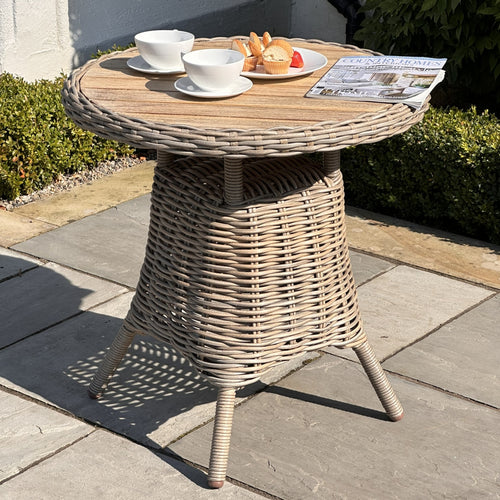 Round outdoor bistro table with a wood top and a brown wicker base, set on a paved area with a couple of cups and a plate on top.