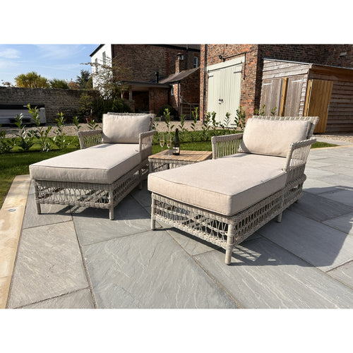 Two beige outdoor sun lounger with cushions and a matching table made of rattan material, placed on a patio.