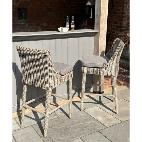 Two beige outdoor bar stools made of woven material with gray cushions, placed on a stone patio.