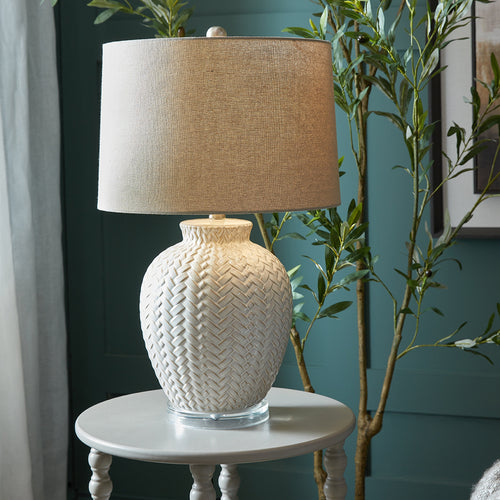 Decorative lamp with textured beige shade and base on a small round table against a teal wall.